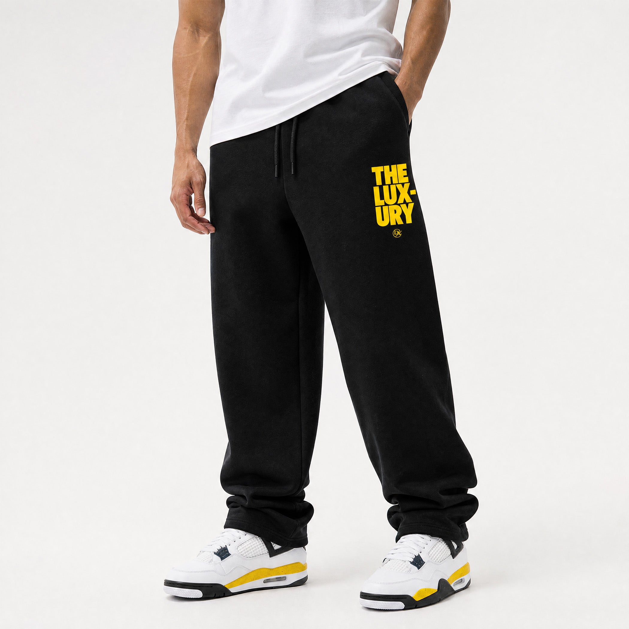 A person wearing black sweatpants with a bold yellow 'THE LUXURY' logo on the left thigh, styled with a white t-shirt and white sneakers with yellow accents, against a clean white studio background.