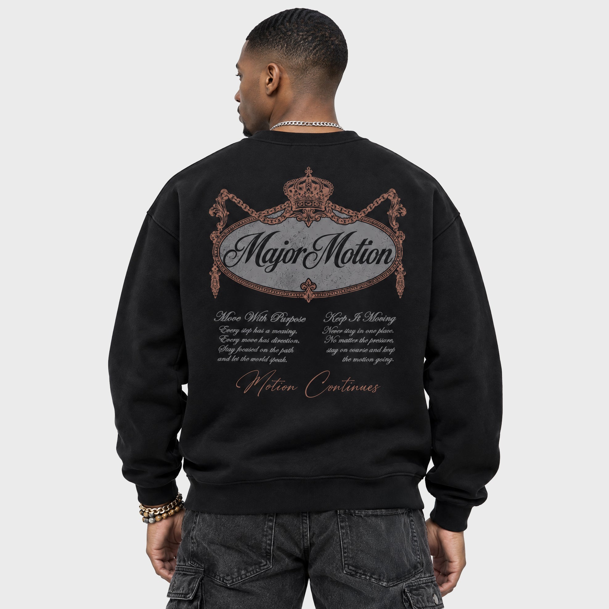 A man viewed from the back wearing a black crewneck sweatshirt with a large graphic print on the back. The graphic features a crown, the words 'Major Motion' in script, decorative flourishes, and smaller white and metallic-looking text below.