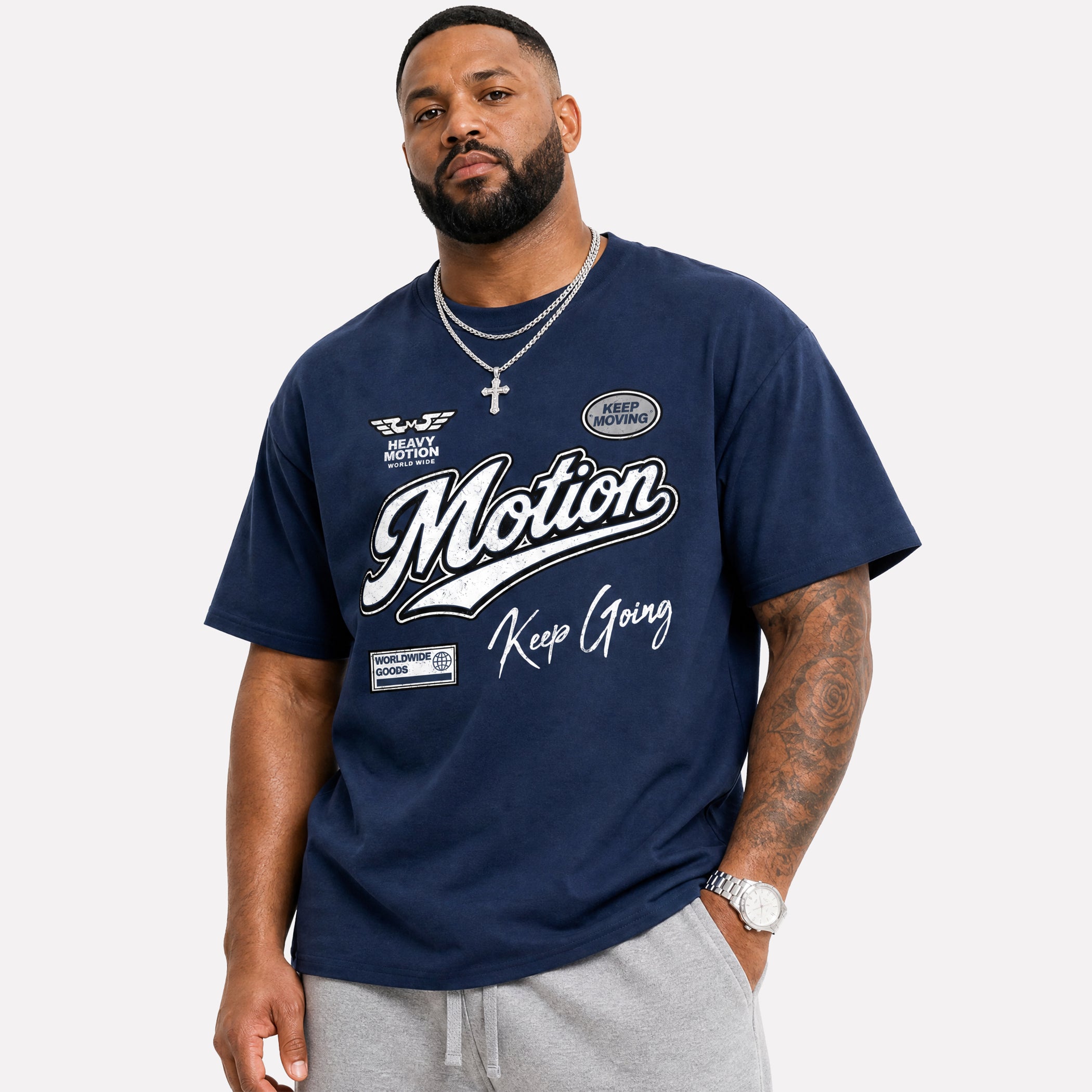 A man wearing a navy blue t-shirt with a white graphic print that reads 'Motion Keep Going' and 'American Motorsport' logos, paired with gray sweatpants against a light gray background.