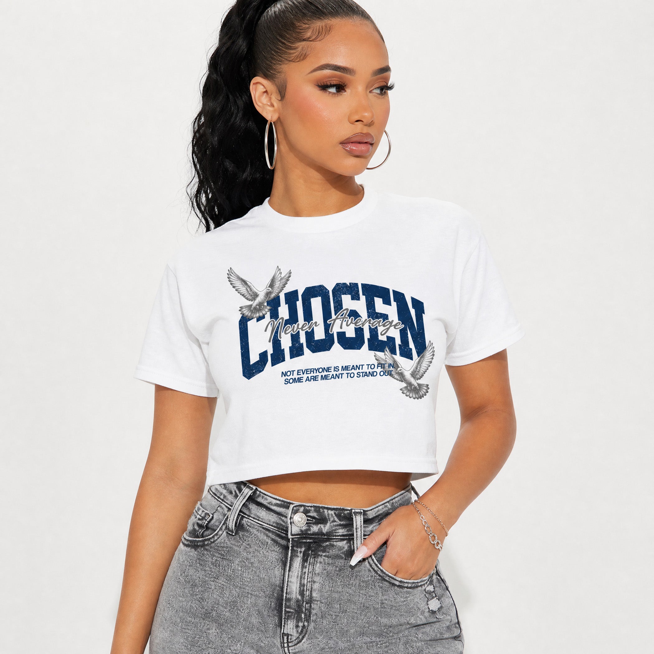 A woman wearing a white cropped graphic t-shirt with 'CHOSEN' printed in navy blue and silver glitter-style lettering, paired with gray denim jeans, against a plain white studio background.
