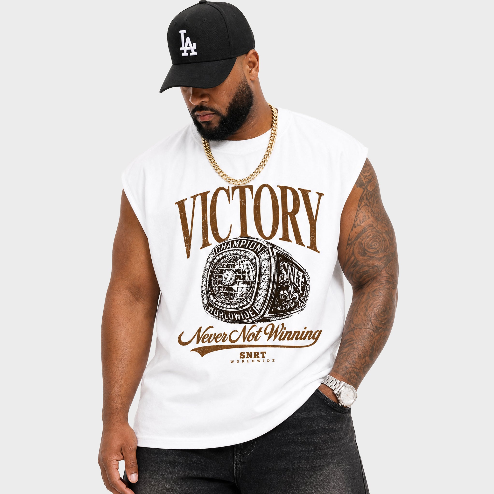 A man wearing a white sleeveless t-shirt with a brown and black championship graphic that reads 'VICTORY Never Not Winning' and 'SNRT APPAREL'. He is also wearing a black Los Angeles Dodgers baseball cap, a gold chain, and dark jeans against a light gray background.