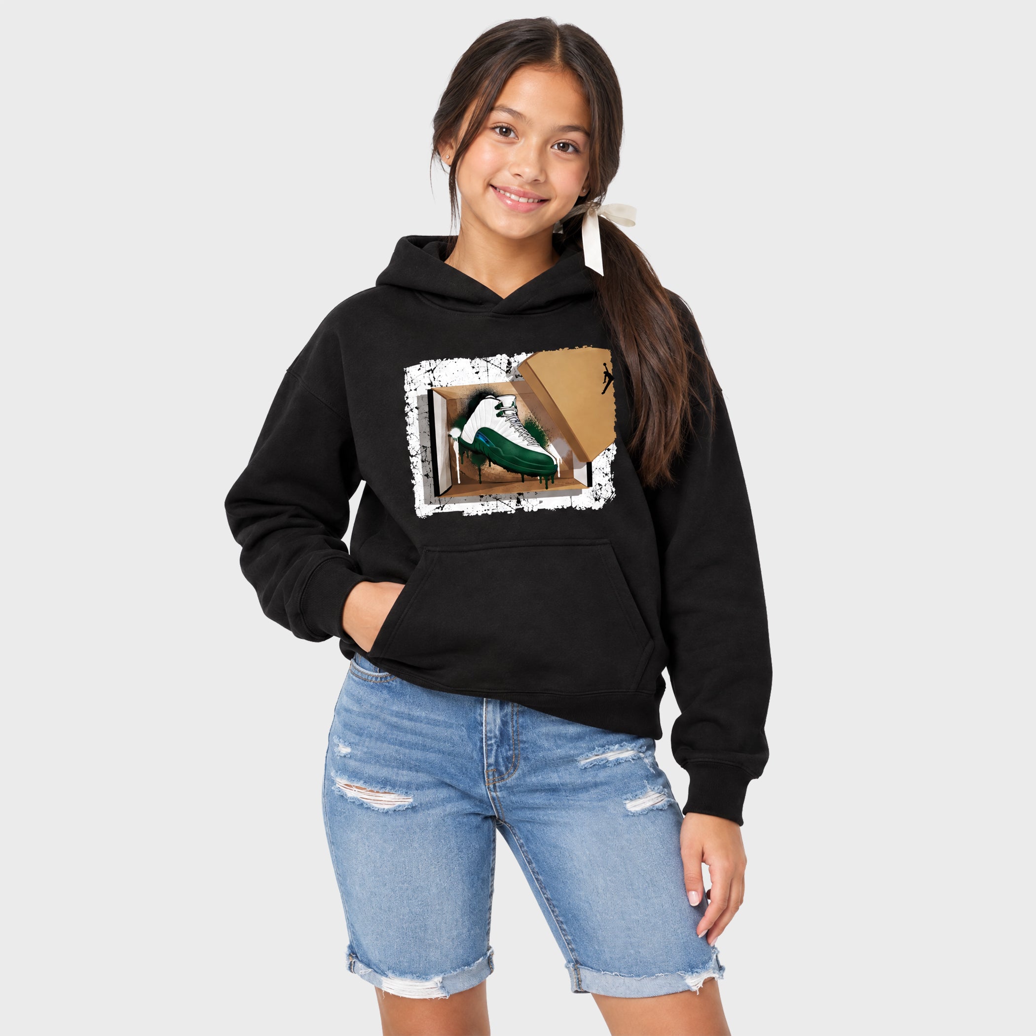 A young girl with dark hair in a ponytail wearing a black hoodie with a graphic print of a green and white sneaker in a cardboard box, paired with light blue denim shorts, against a light gray background.