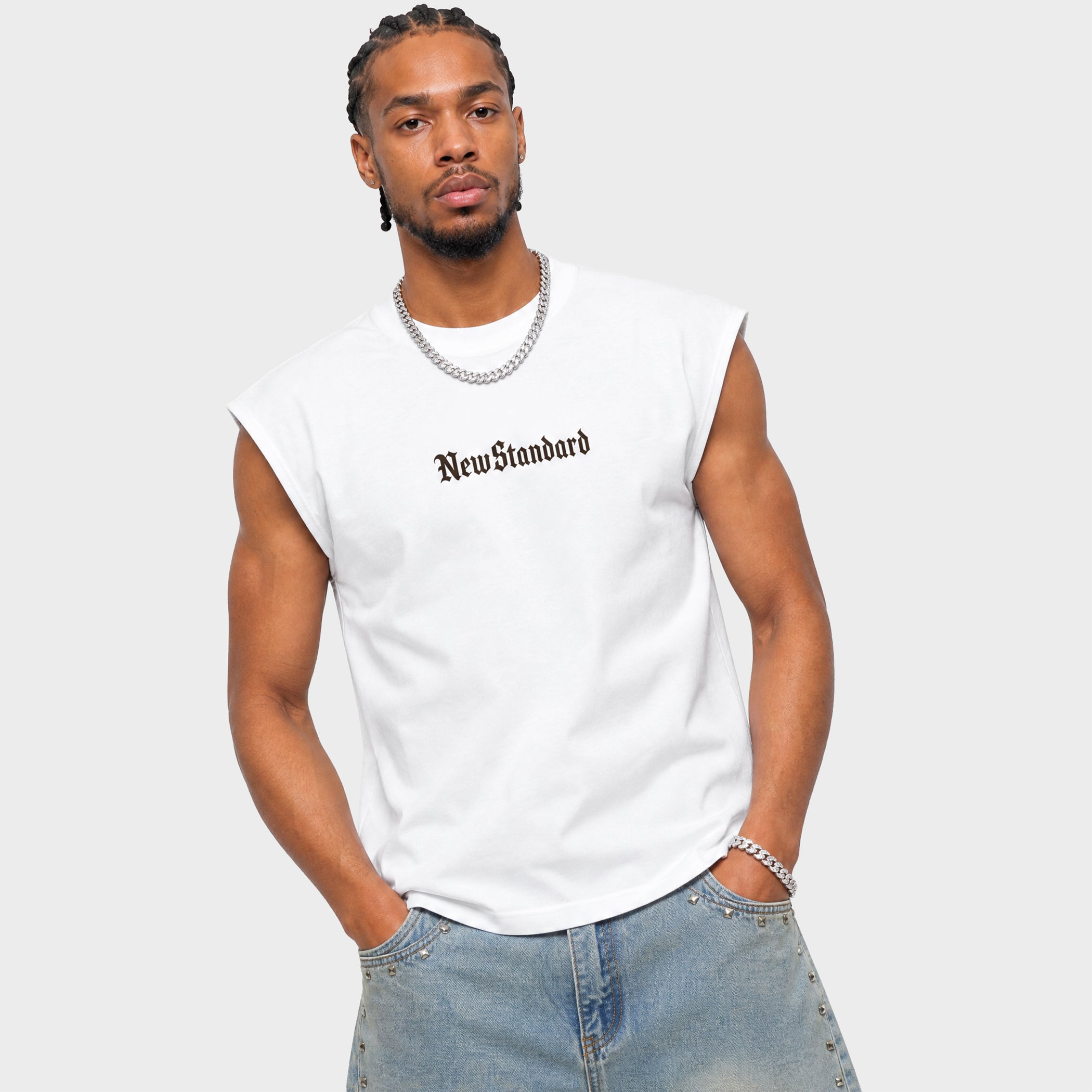 A man wearing a white sleeveless muscle t-shirt with 'NewStandard' printed in black Gothic font on the chest, paired with light blue jeans and silver jewelry, against a light gray background.