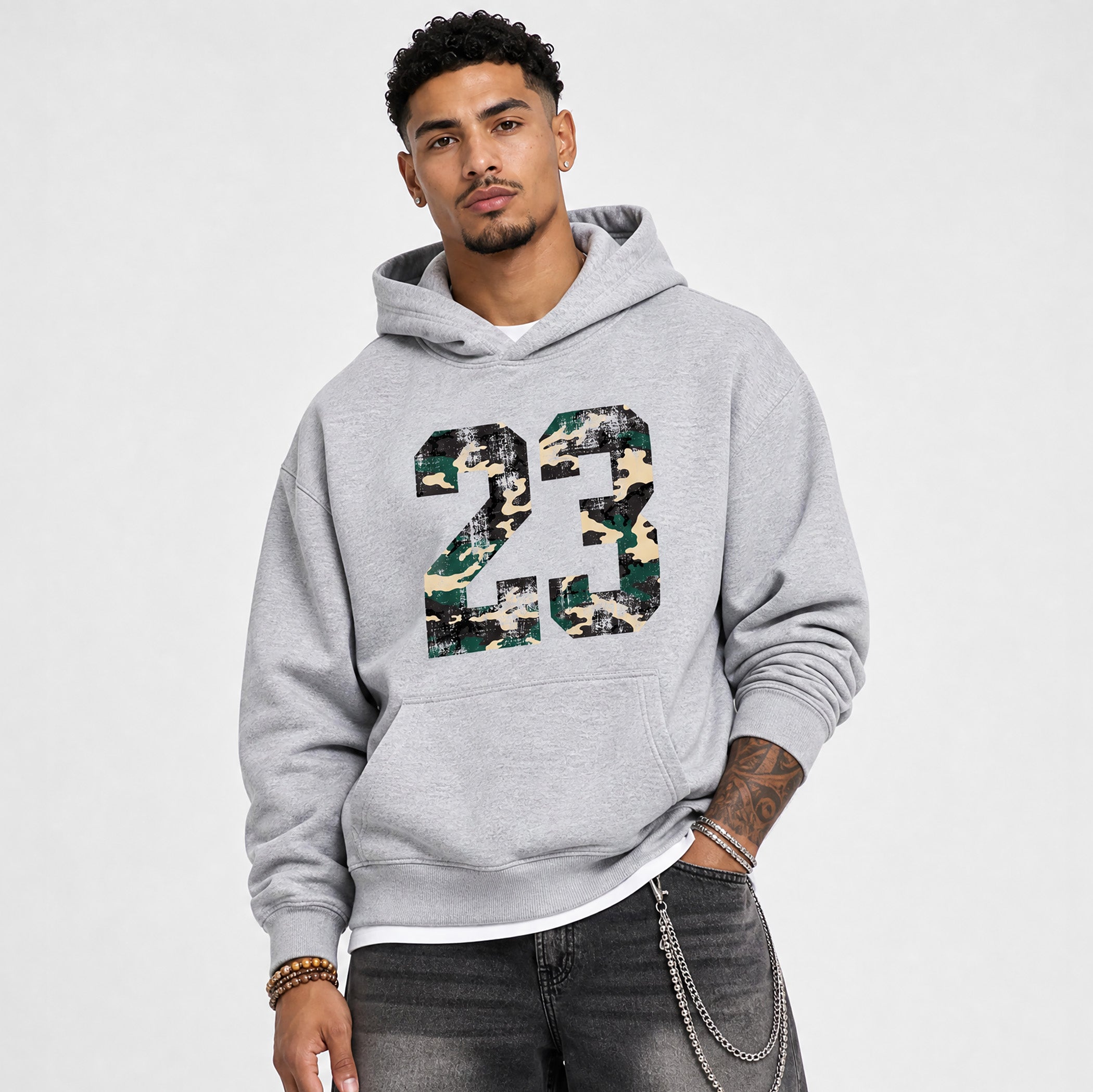 A man wearing a light gray pullover hoodie with a large '23' graphic on the front filled with a green, brown, and beige camouflage pattern. He is also wearing dark gray jeans with silver chain accessories.