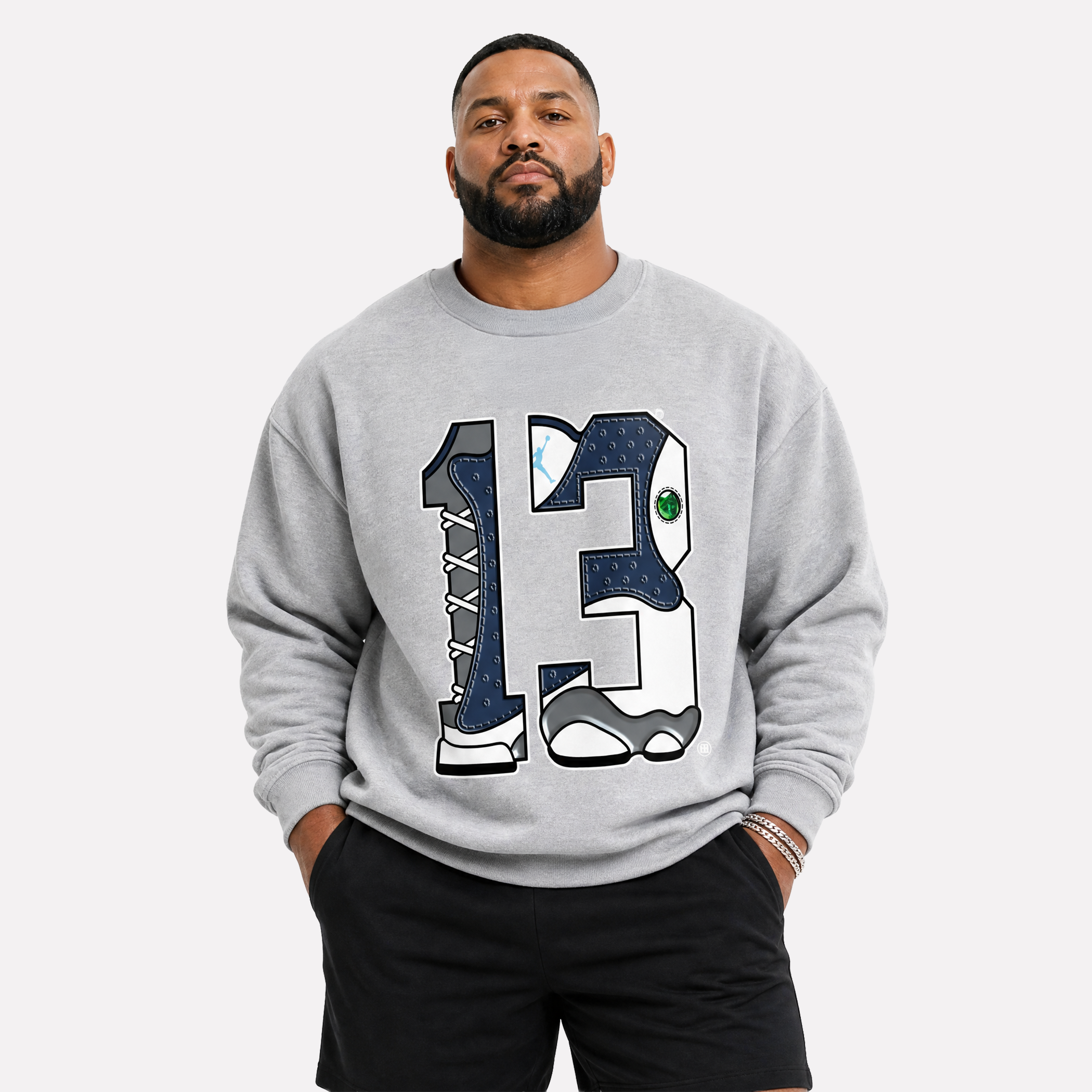 A man wearing a light gray crewneck sweatshirt with a large graphic of the number 13 filled with sneaker designs, paired with black shorts, against a light gray background.