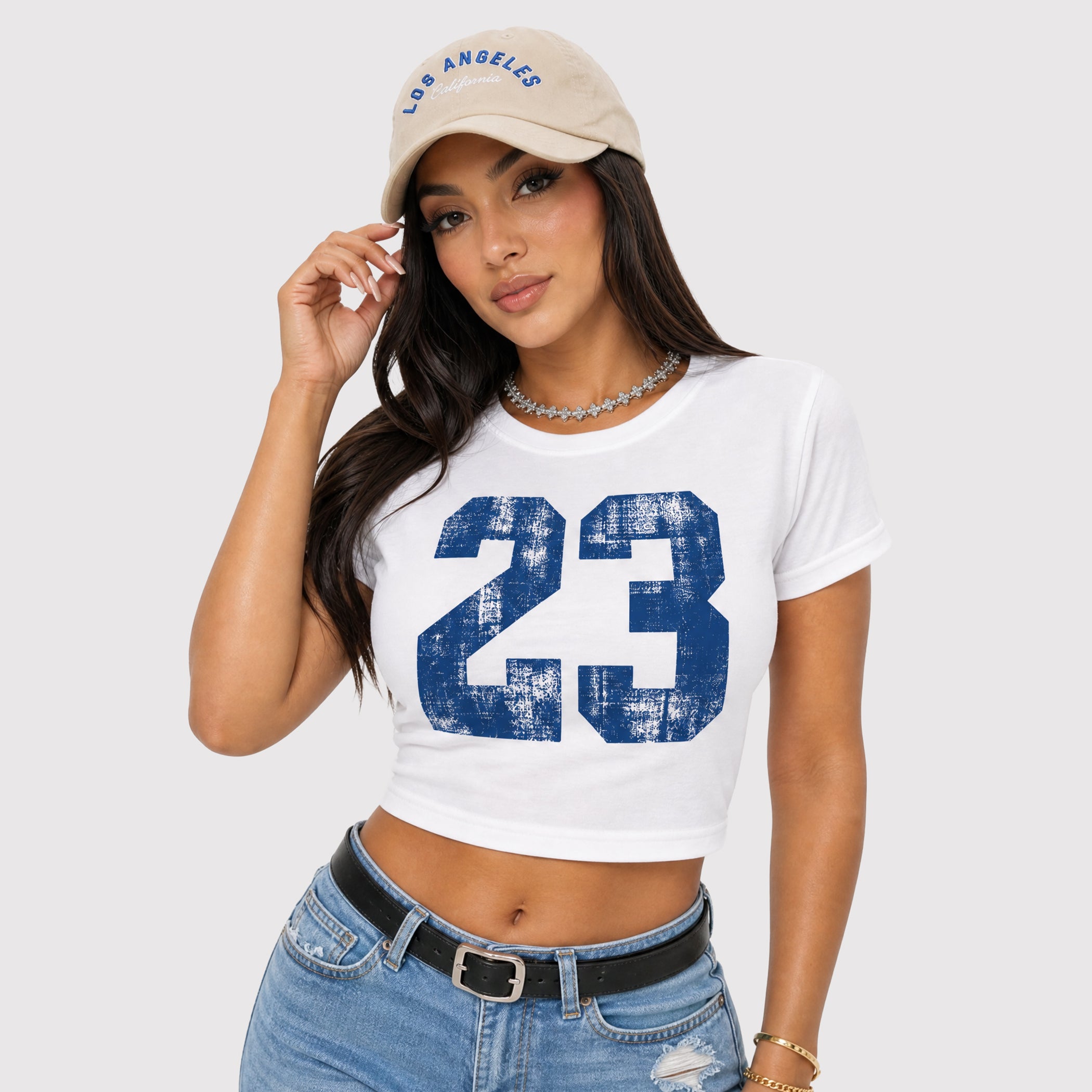 A woman wearing a white cropped t-shirt with a distressed blue number 23 graphic, paired with a beige Los Angeles baseball cap and blue jeans, against a light gray background.