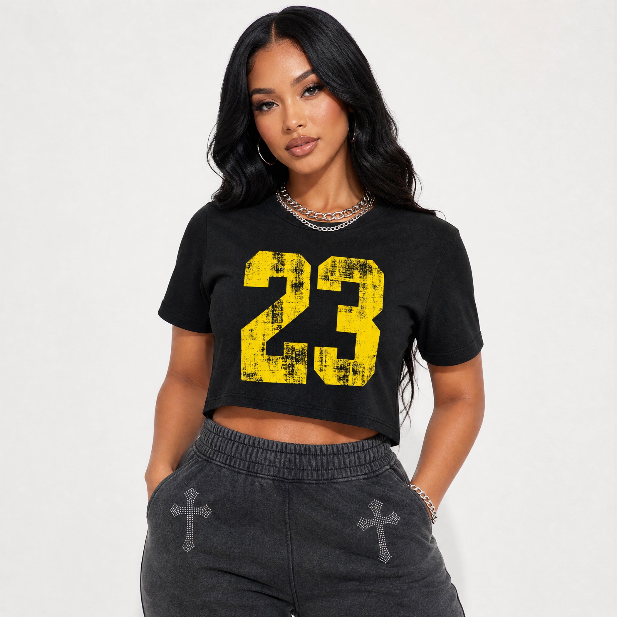 A woman modeling a black cropped t-shirt with a large distressed yellow number 23 graphic on the front, paired with dark gray sweatpants, against a plain light gray background.