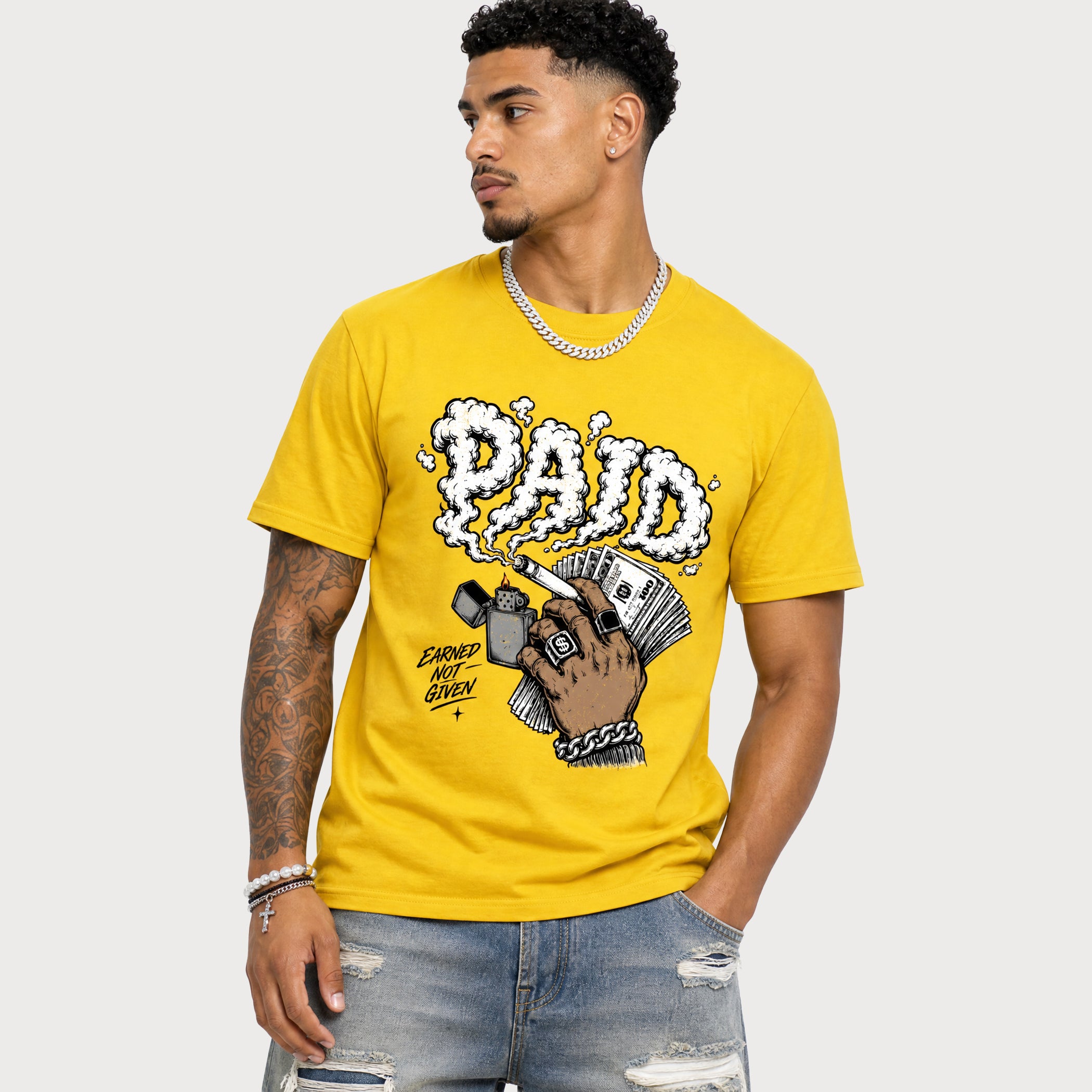 A man wearing a bright yellow graphic t-shirt with a 'PAID' hand illustration, paired with distressed blue jeans and a silver chain necklace, set against a plain light gray background.
