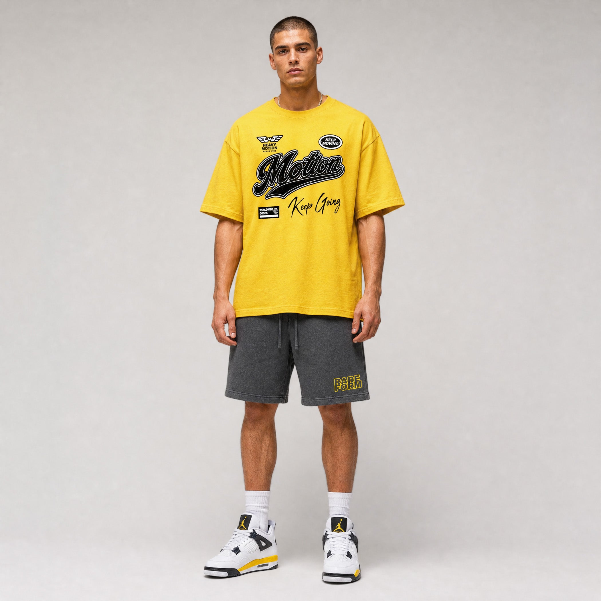 A male model stands against a light gray studio background wearing an oversized bright yellow graphic T-shirt with dark lettering, dark gray shorts with yellow text, white socks, and white sneakers with yellow and black accents.