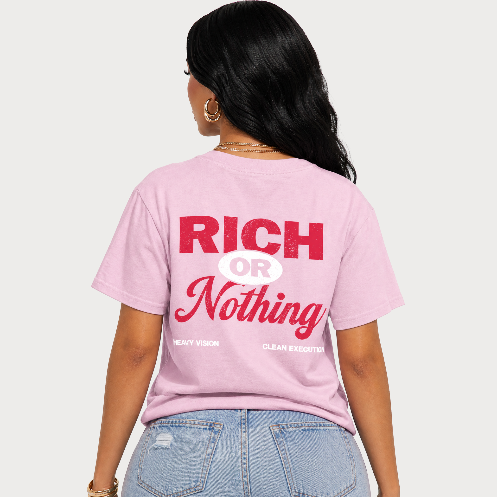 A woman shown from the back wearing a light pink graphic t-shirt with the text 'RICH OR Nothing' in red and white, paired with light blue jeans, against a solid light gray background.