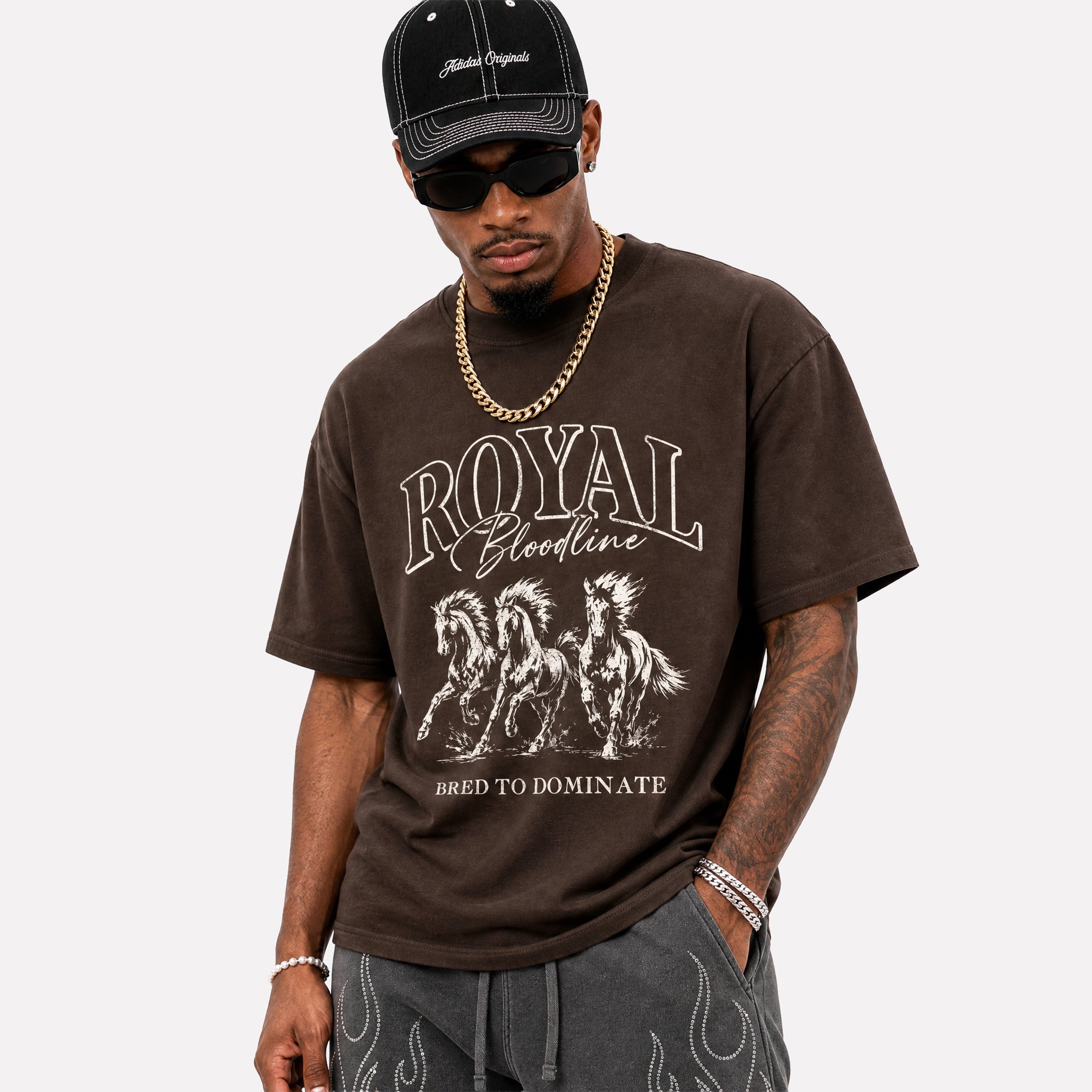 A man wearing a dark brown graphic t-shirt with 'ROYAL Bloodline' and horse illustrations, paired with a black cap, sunglasses, and a gold chain, against a light gray background.