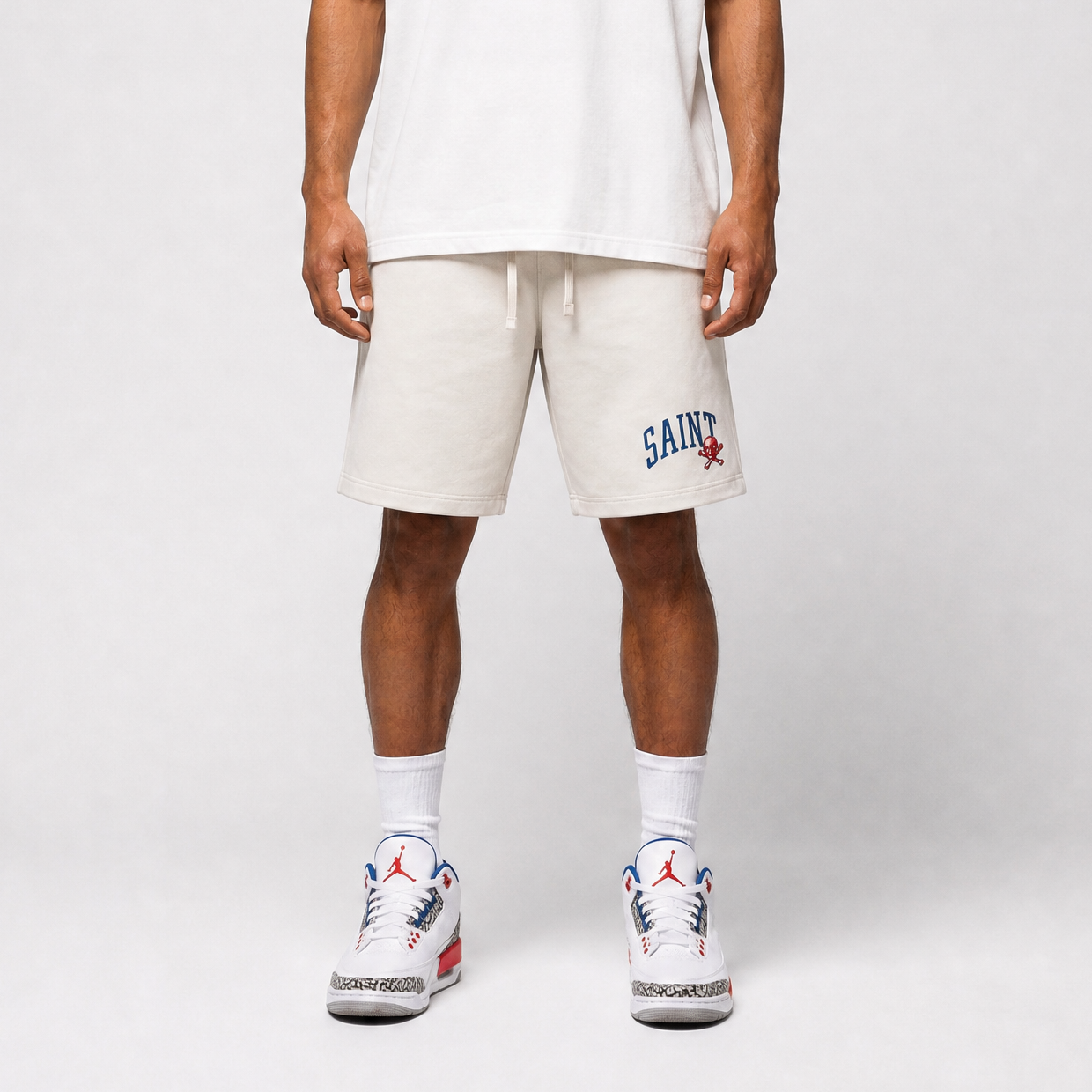 Person wearing white shorts with 'SAINT' branding and matching sneakers on a plain background