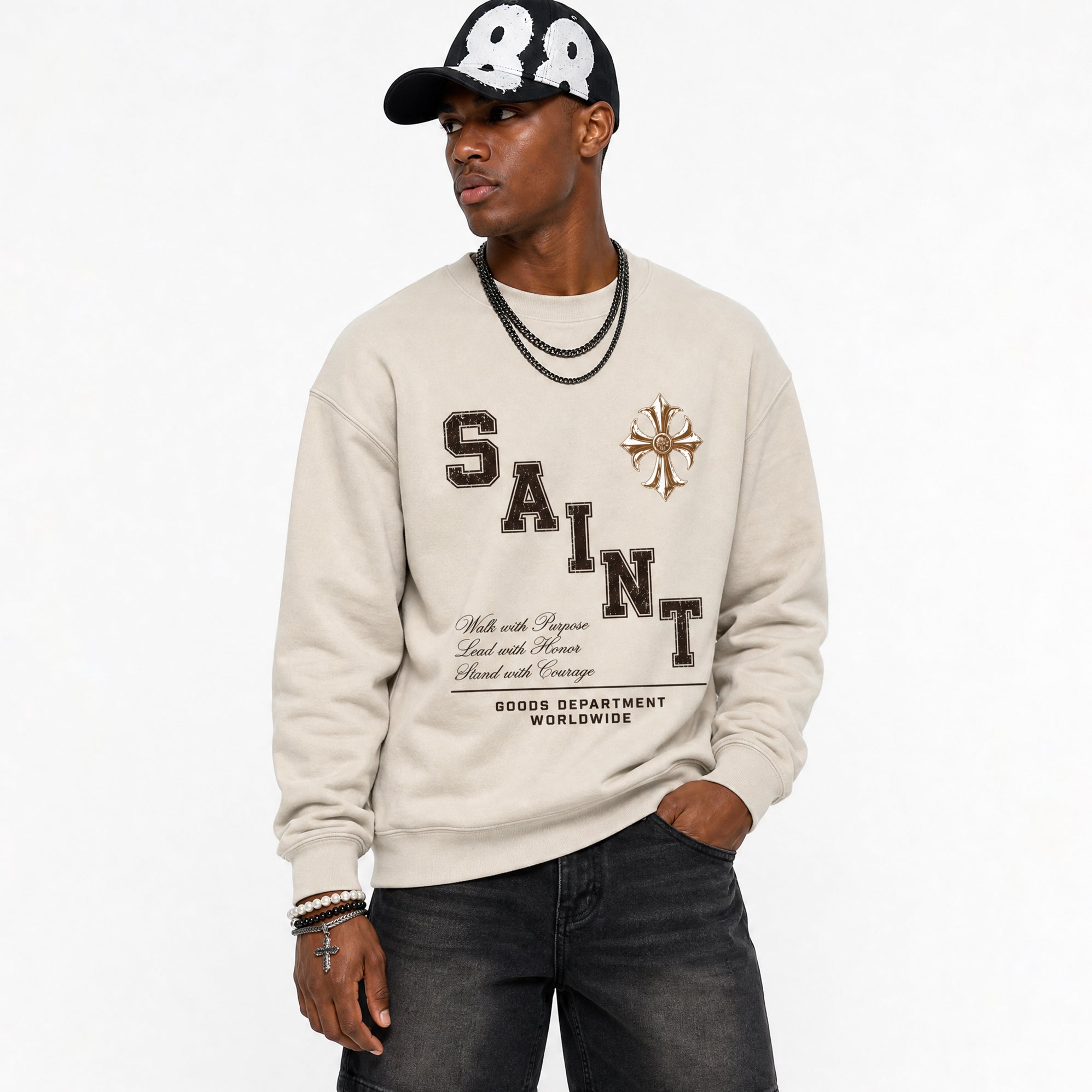 A man wearing a beige Saint Laurent sweatshirt with a brown graphic print, a black baseball cap with white numbers, and dark gray jeans, photographed against a plain white background.