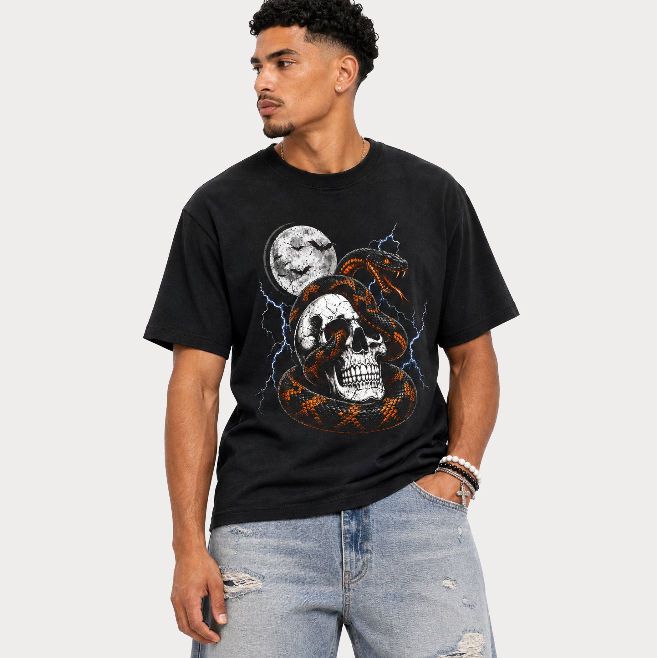 A man wearing a black graphic t-shirt with a skull, snake, and moon design, paired with light-wash distressed jeans, set against a plain light gray background.