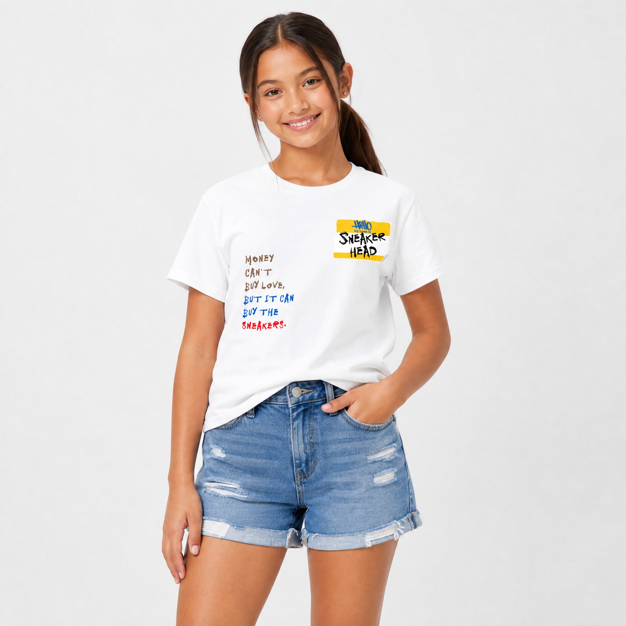 A young girl with her hair in a ponytail models a white graphic t-shirt and distressed denim shorts against a light gray studio background. The t-shirt features a yellow 'SWEETENER HEAD' logo and colorful handwritten text on the left chest.