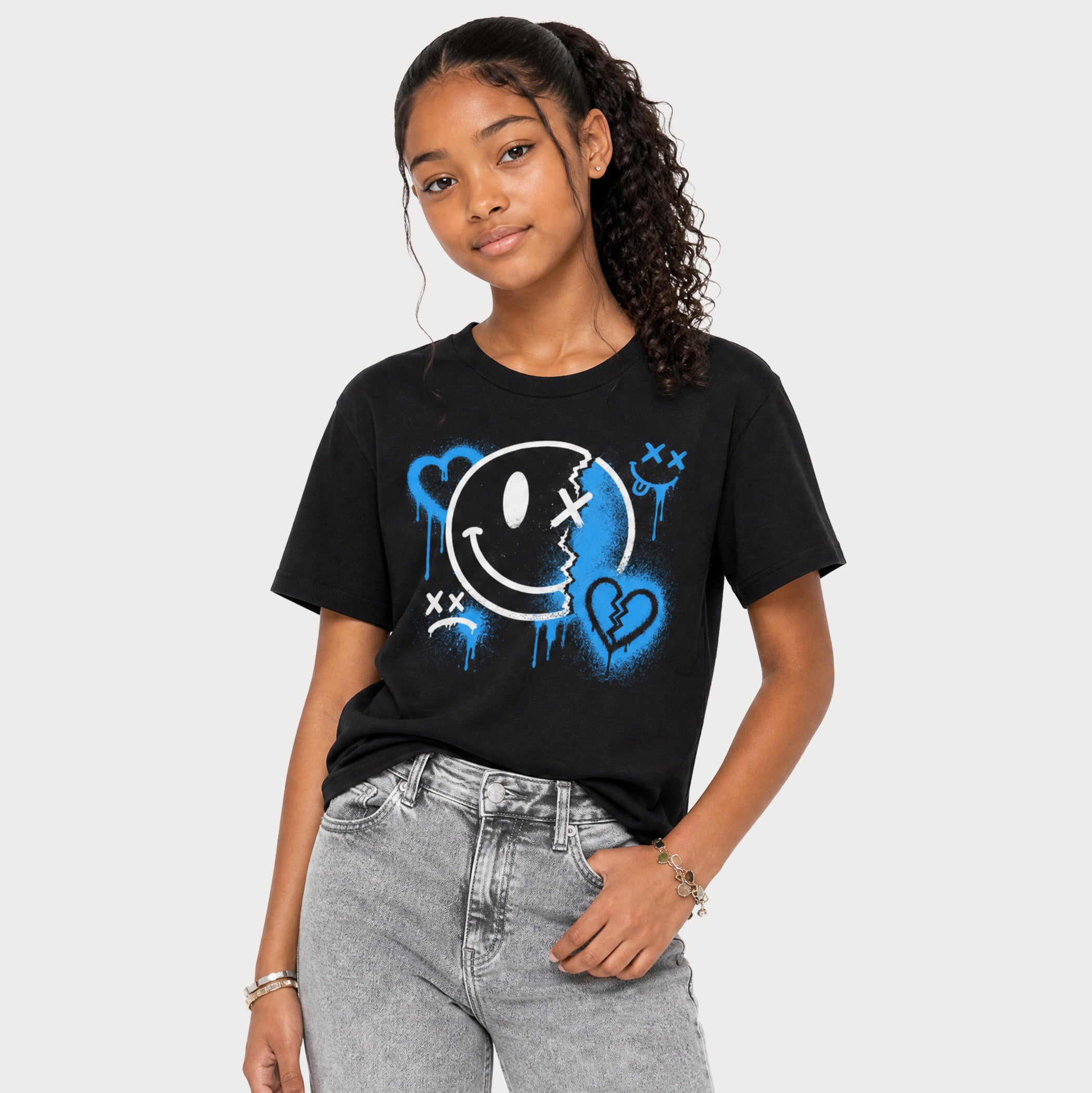 A woman modeling a black graphic t-shirt with a blue and white graffiti-style smiley face design, paired with light-wash jeans, against a solid light gray background.