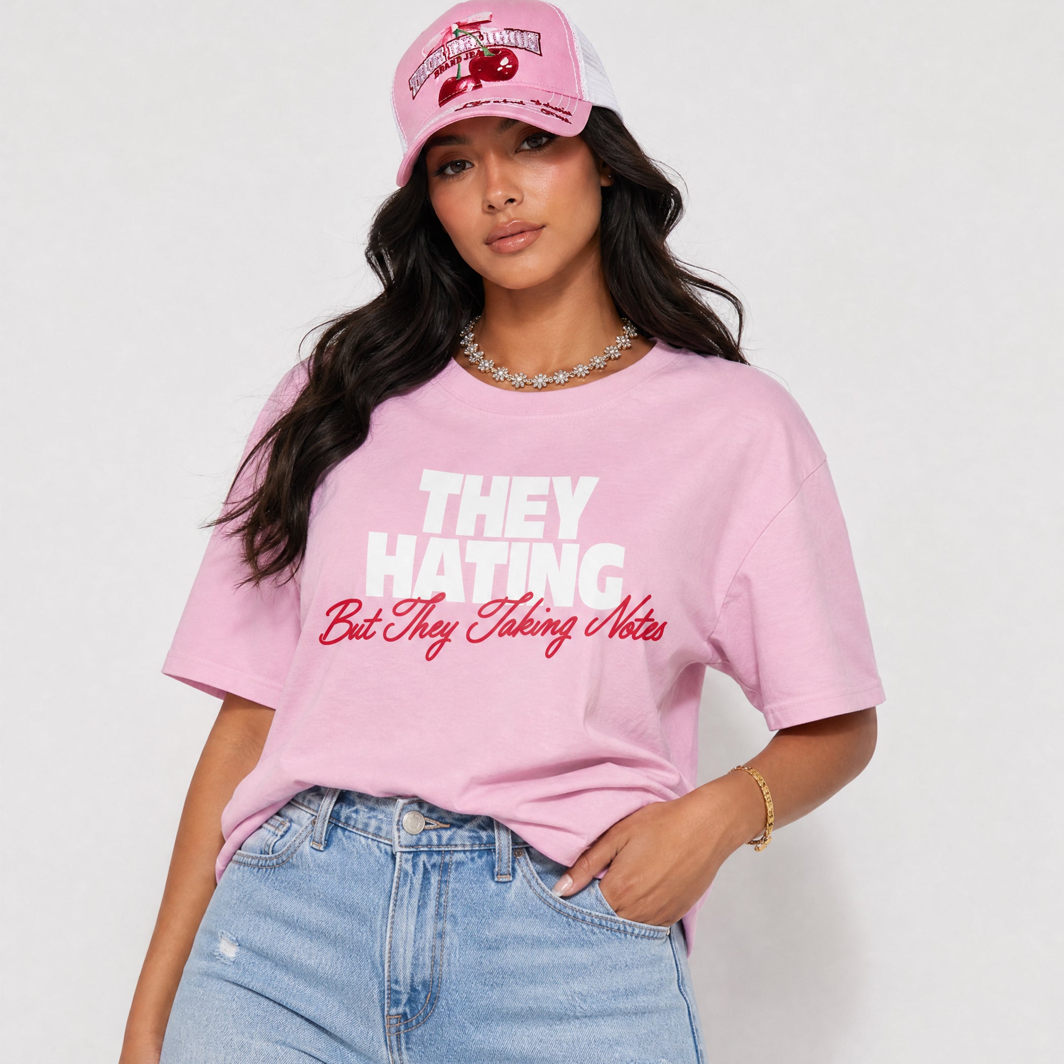 A woman wearing a light pink graphic t-shirt with the text 'THEY HATING But They Taking Notes' in white and red, paired with a matching pink cap and light blue jeans, against a plain light gray background.