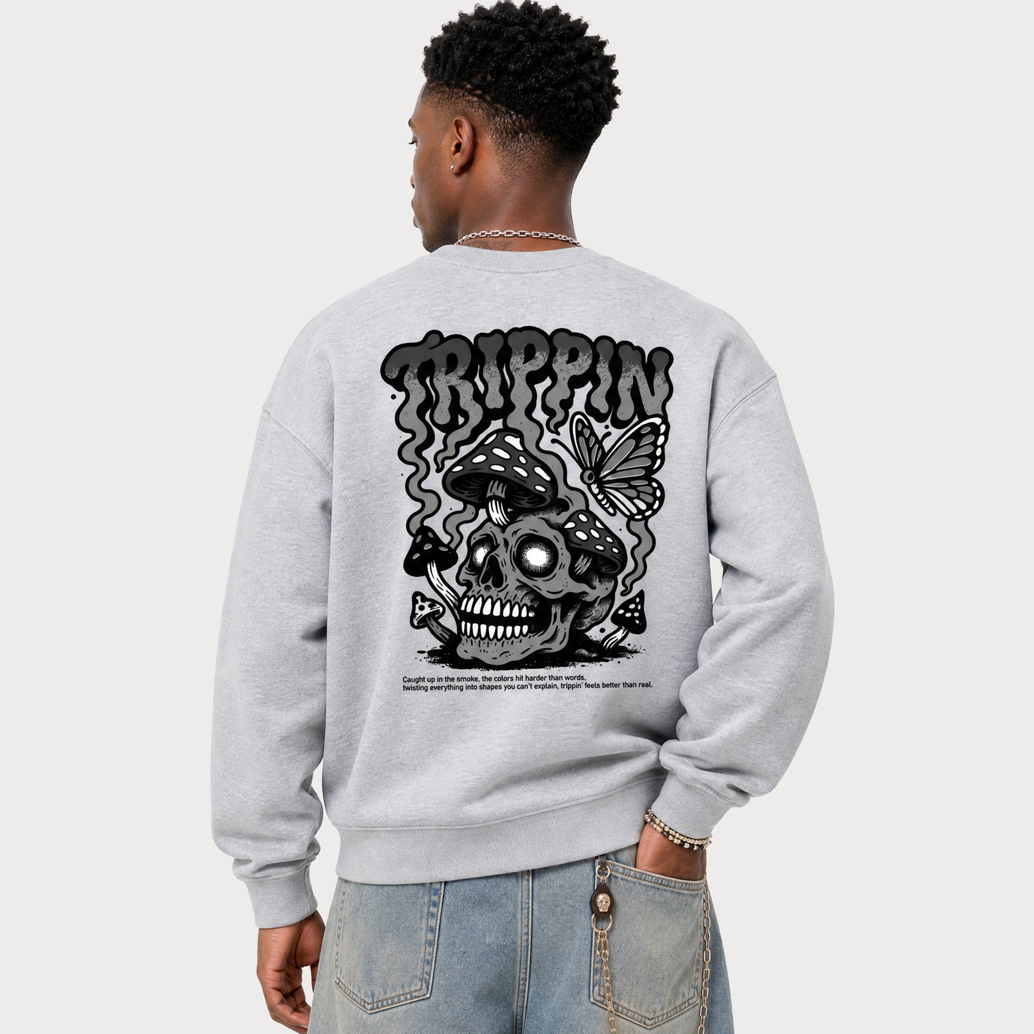 A man shown from the back wearing a light gray crewneck sweatshirt with a large black graphic on the back featuring a skull, butterfly, and the word 'TRIPPIN'.