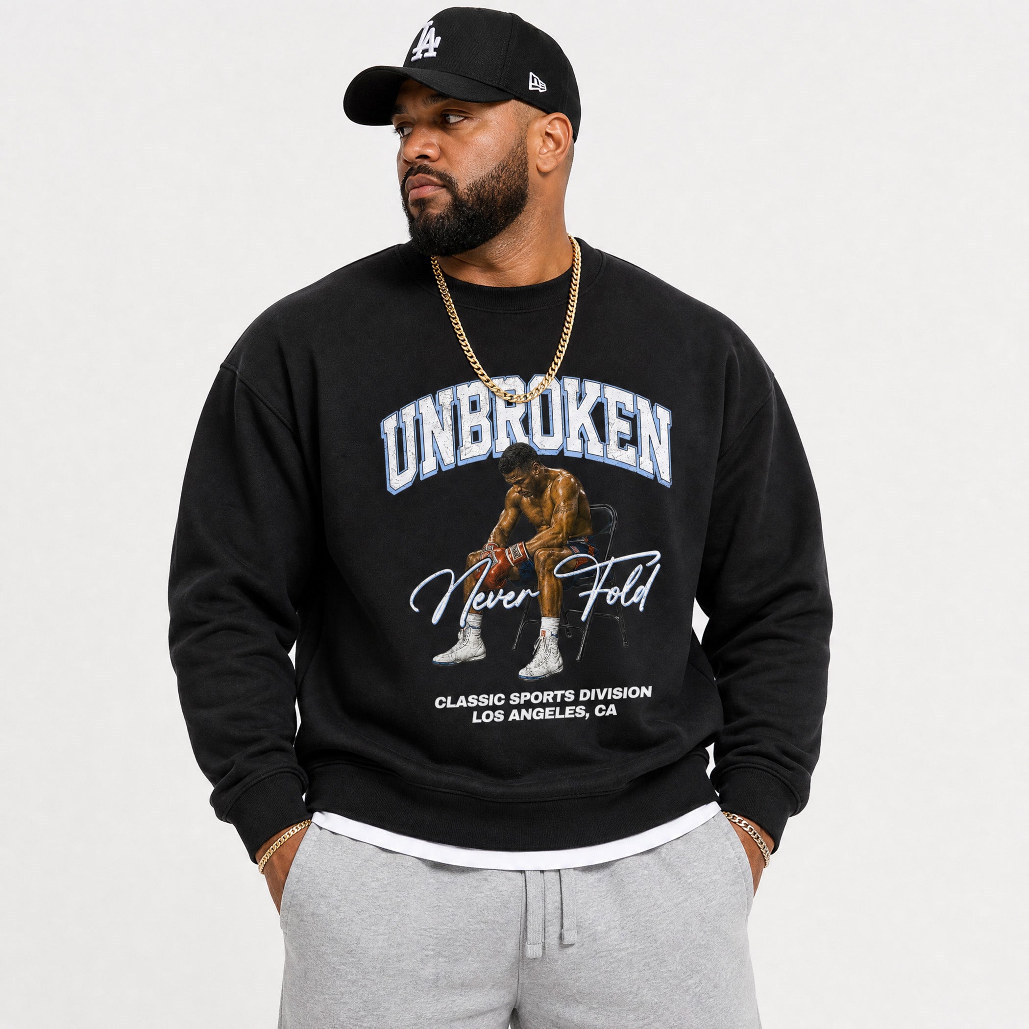 A man wearing a black crewneck sweatshirt with a graphic of Kobe Bryant and the text 'UNBROKEN Never Fold', paired with a black Los Angeles Dodgers cap, gold chain, and gray sweatpants, against a light gray background.