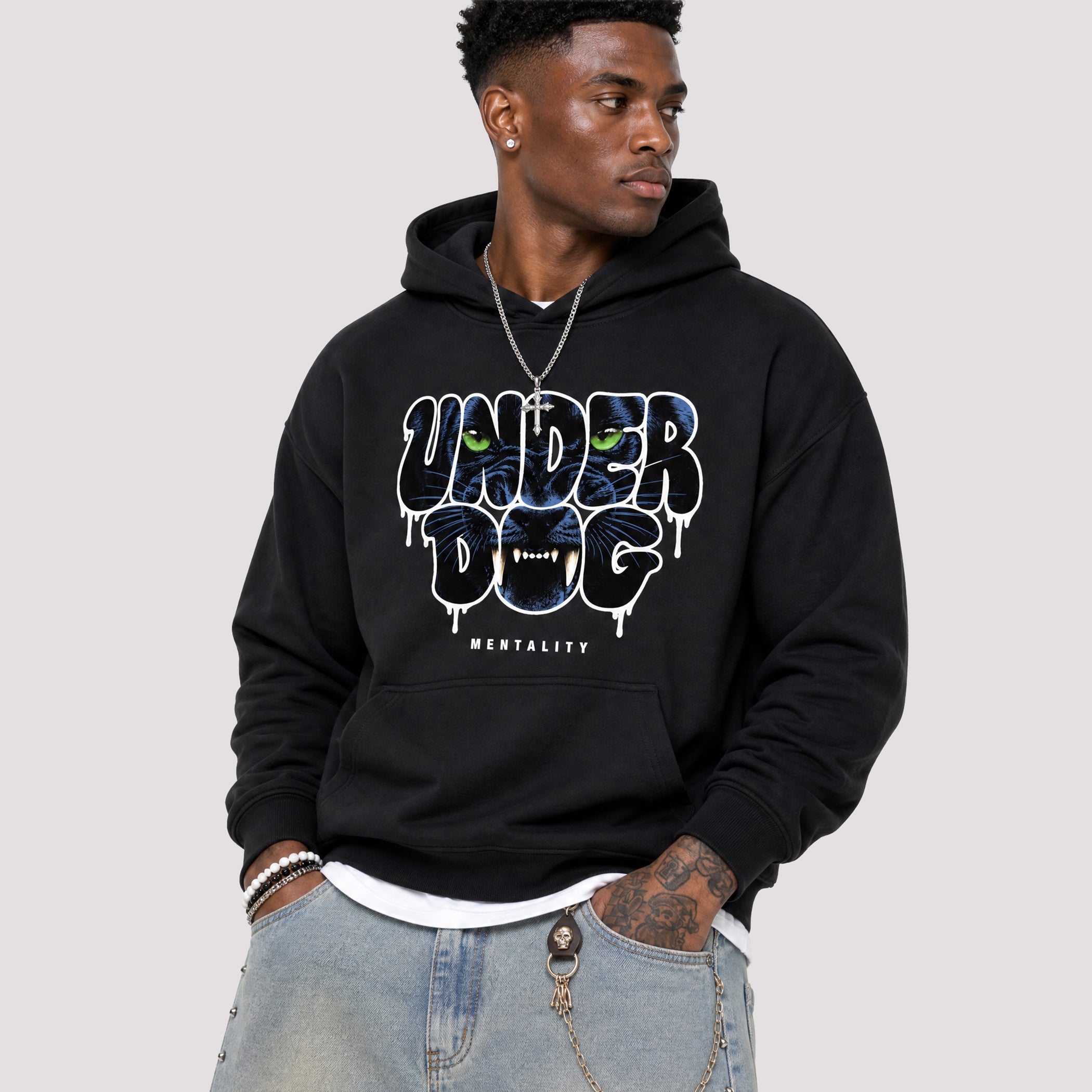 A man wearing a black pullover hoodie with a large graphic on the chest that reads 'UNDER DOG' in dripping white letters with blue and green eyes, and 'MENTALITY' below it, paired with light blue jeans against a solid light gray background.