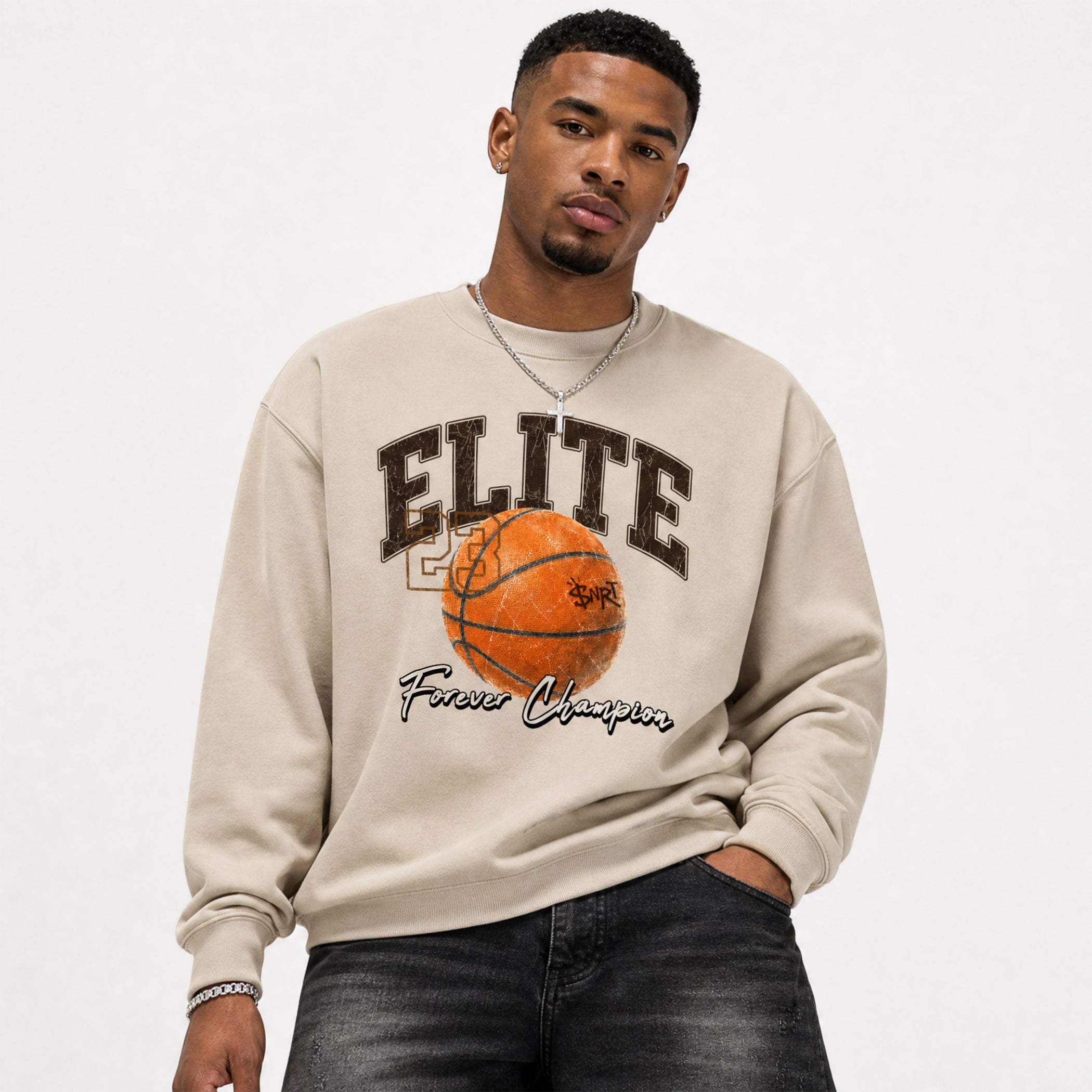 A man wearing a beige crewneck sweatshirt with a graphic print of the word 'ELITE' in dark brown, an orange basketball, and the words 'Forever Champion' in black script. He is also wearing dark gray jeans and a silver chain necklace, photographed against a plain light gray background.